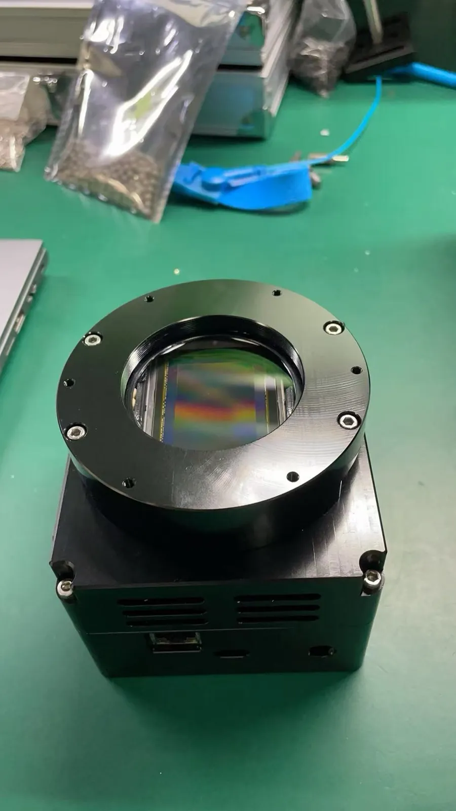 A black engineering-prototype camera body with a large round sensor window on a workbench
